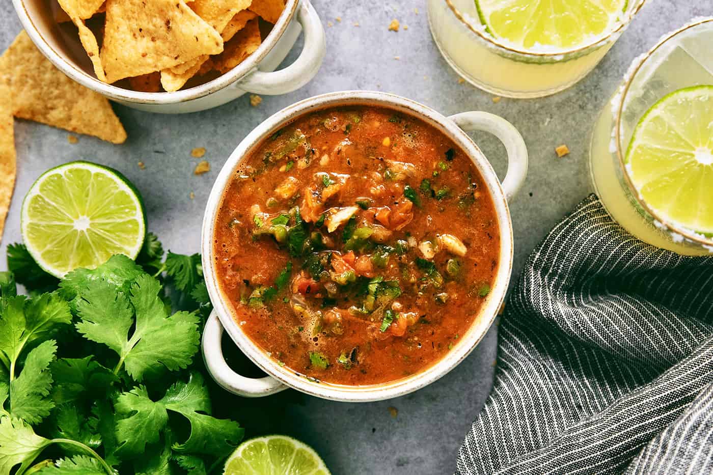 A white handled bowl holds warm salsa with a bowl of tortilla chips next to it along with limes.