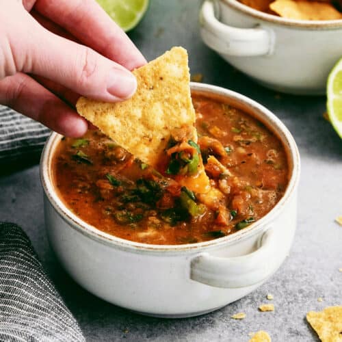 A hand dips a tortilla chip into a bowl of warm salsa.
