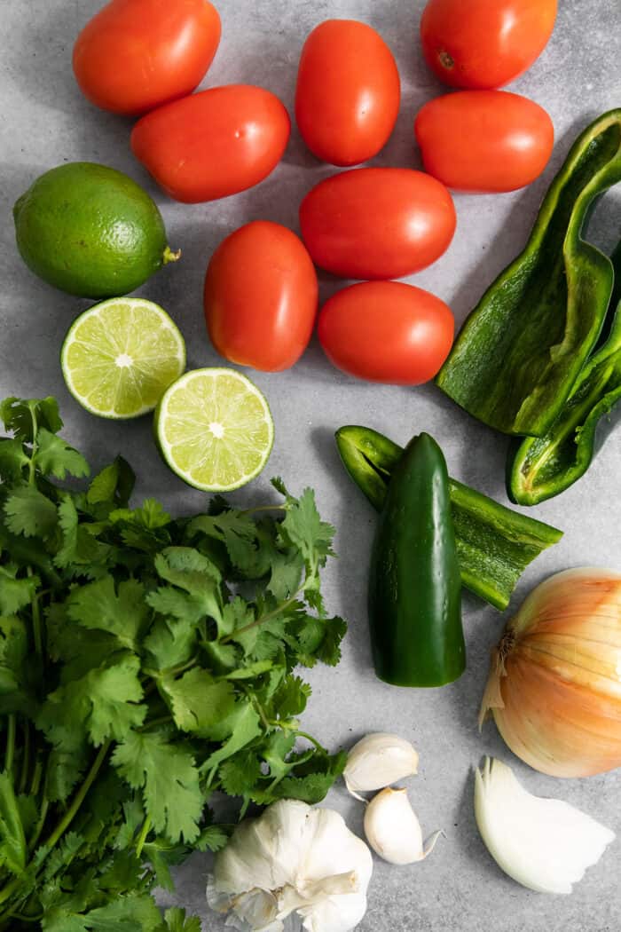 This Warm Salsa recipe is perfectly spiced and juicy, with a pop of lime juice for sweetness. Enjoy it with tacos, quesadillas, & chips. The ingredients for salsa are shown including tomatoes, limes, green peppers, onion, and garlic.