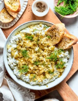 A bowl of dill pickle dip is topped with potato chips and dill and served with bread.