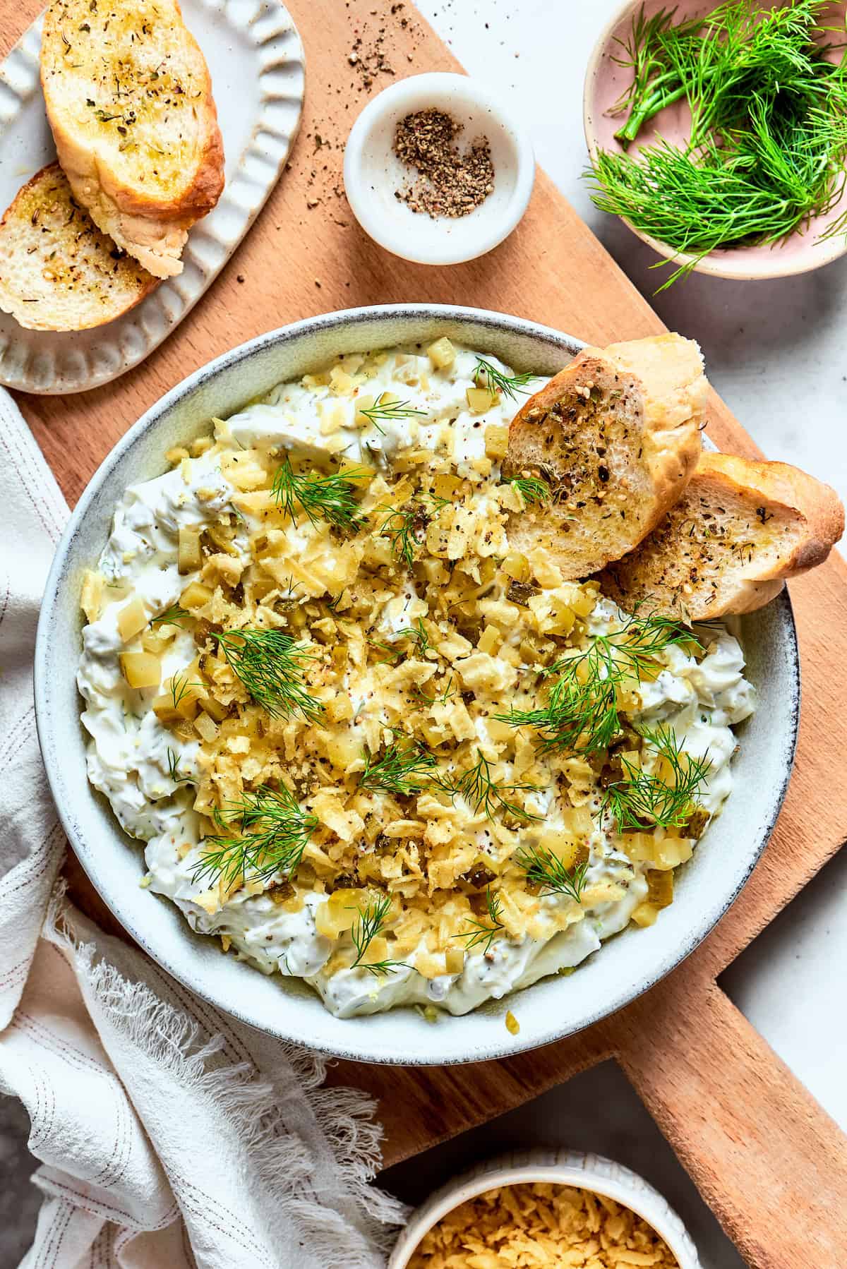 This cold and creamy Dill Pickle Dip is packed with crunchy pickles and a mix of cream cheese and sour cream. A bowl of dill pickle dip is topped with potato chips and dill and served with bread.