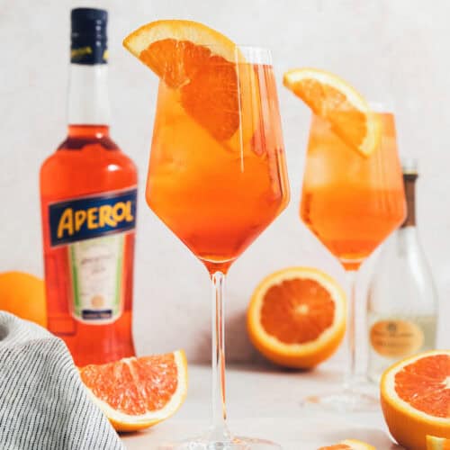 Glasses of Aperol spritz are surrounded with orange wedges next to a bottle of Aperol.