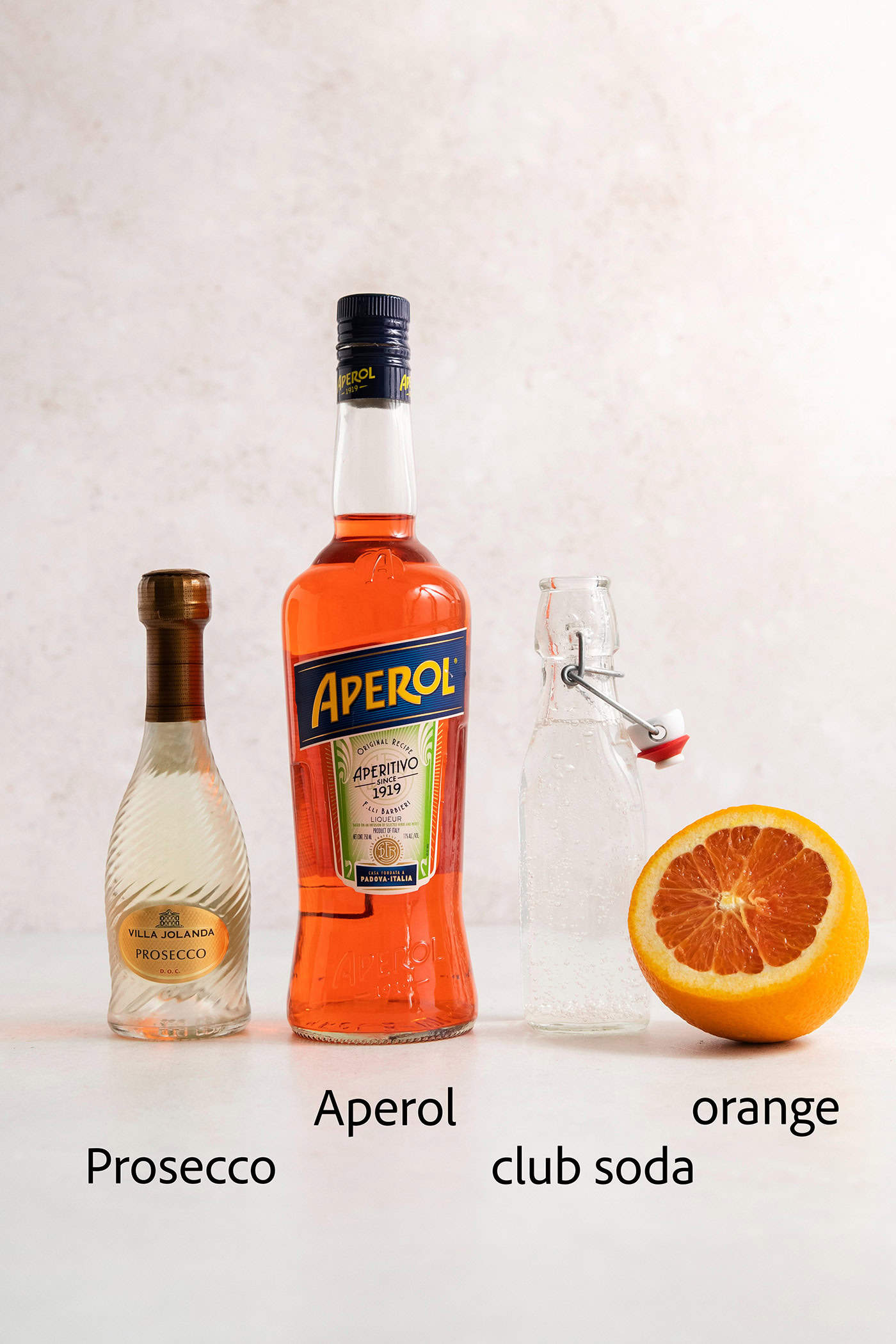 This classic Aperol spritz recipe is my take on an Italian aperitivo and makes a refreshing cocktail out of just four ingredients. Aperol spritz ingredients are shown and labelled: a bottle of prosecco, a bottle of Aperol, club soda, an orange.