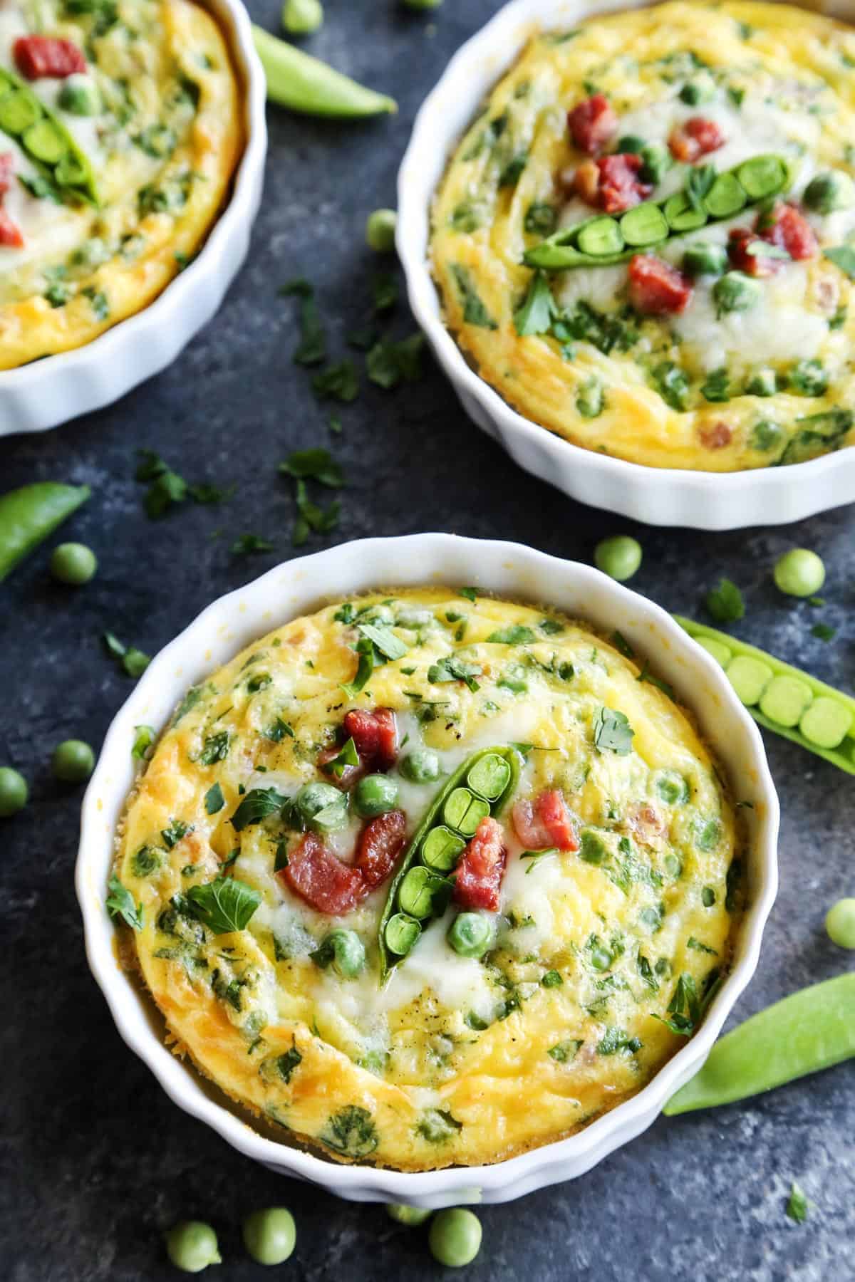 Soft and savory Baked Eggs are a welcome addition to the brunch table. Loaded with salty pancetta, melted provolone, and peas. Three ramekins of baked eggs are topped with peas.