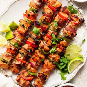 Four grilled charmoula pork kabobs on a plate with lime wedges.