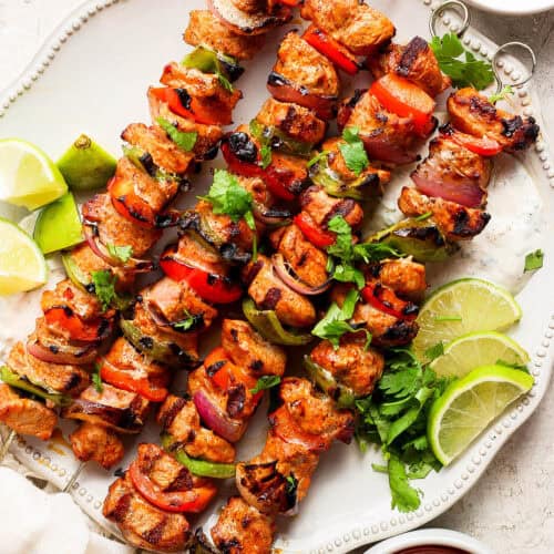 Four grilled charmoula pork kabobs on a plate with lime wedges.
