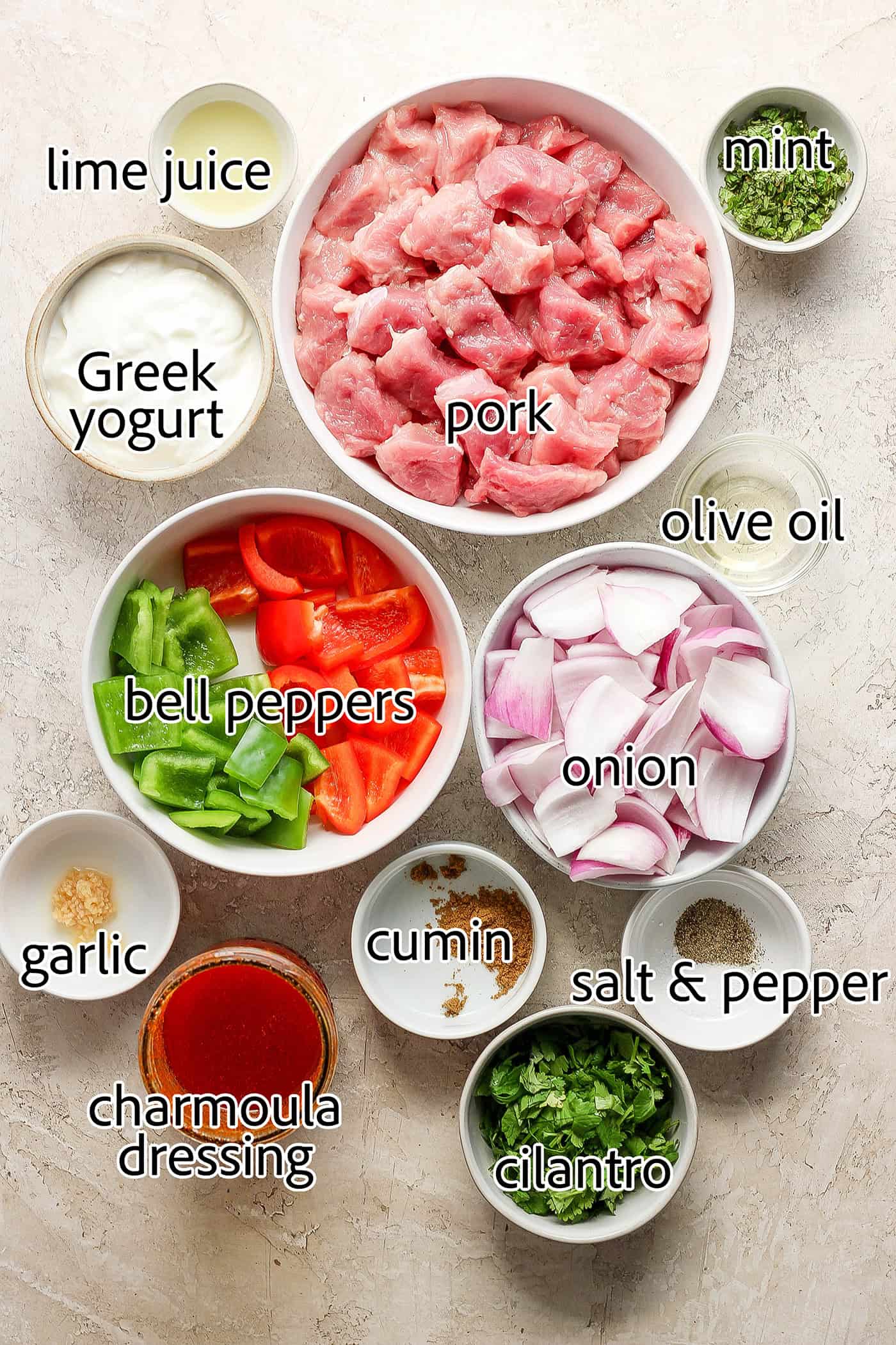 The ingredients for charmoula pork kabobs are shown labelled and portioned out: Greek yogurt, mint, pork, bell peppers, onion, cumin, garlic, charmoula, salt and pepper, olive oil.