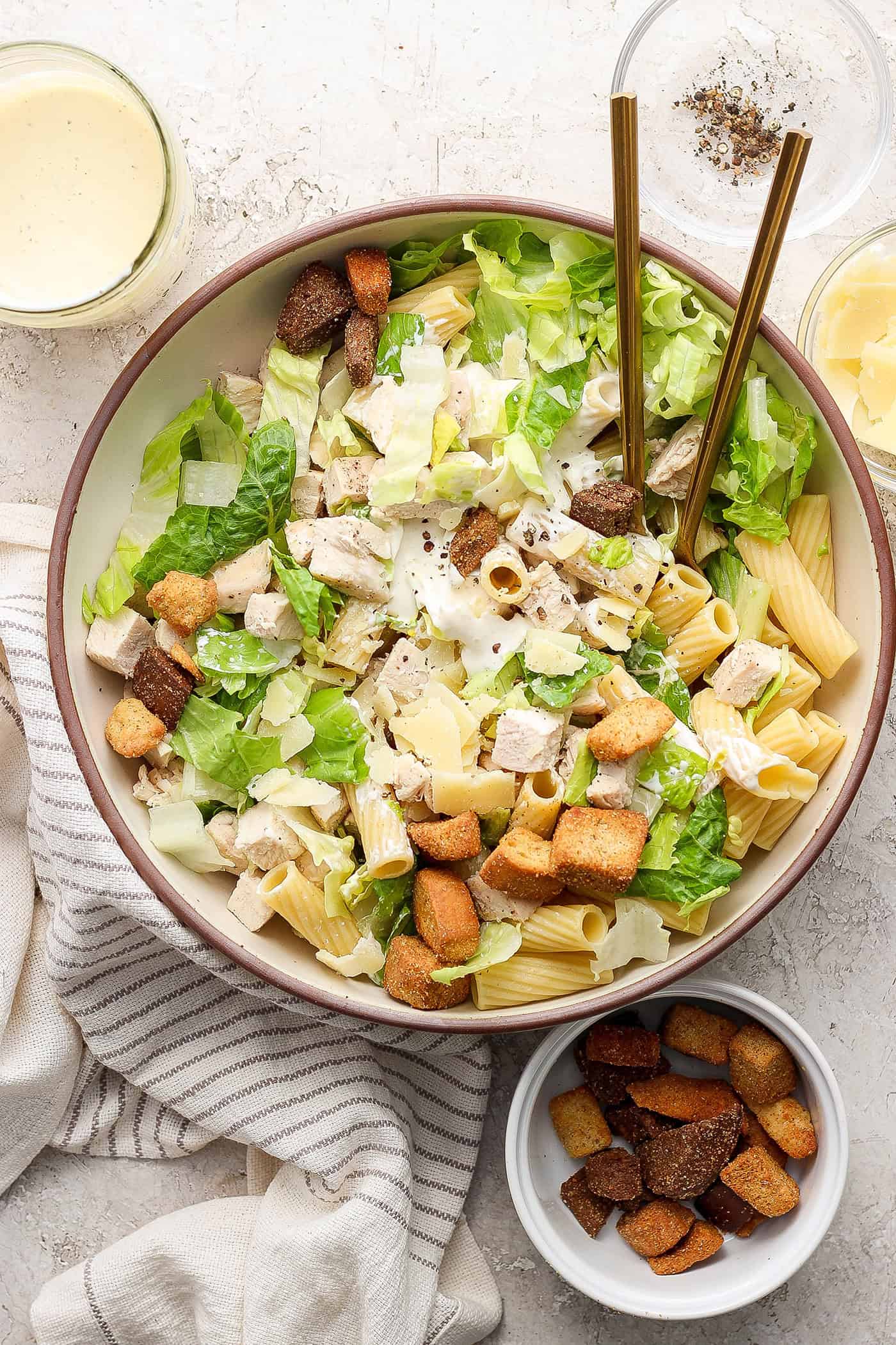 A big bowl of Caesar chicken pasta salad with serving utensils.