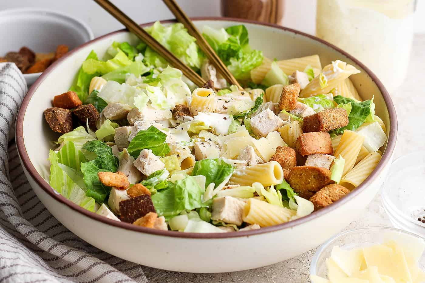 A big bowl of Caesar chicken pasta salad with serving utensils.