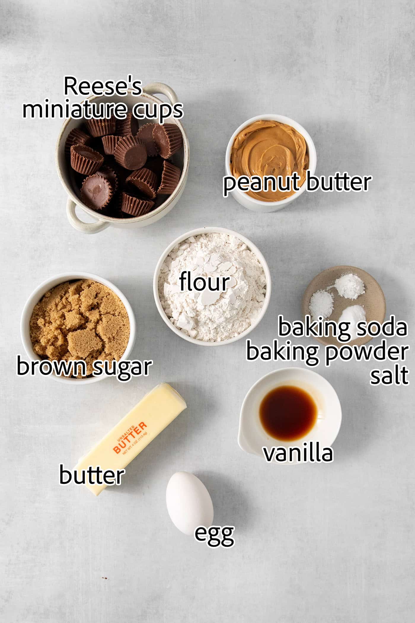 These Peanut Butter Cup Cookies feature soft and chewy peanut butter cookies stuffed with a gooey, chocolatey Reese's cup. Ingredients to make peanut butter cup cookies are shown labelled and portioned out: flour, vanilla, peanut butter, egg, butter, baking powder, baking soda, salt, brown sugar, mini peanut butter cups.