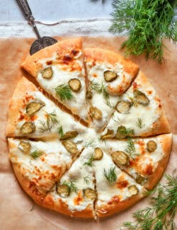 A whole pickle pizza shows it topped with white sauce, pickles, and dill.