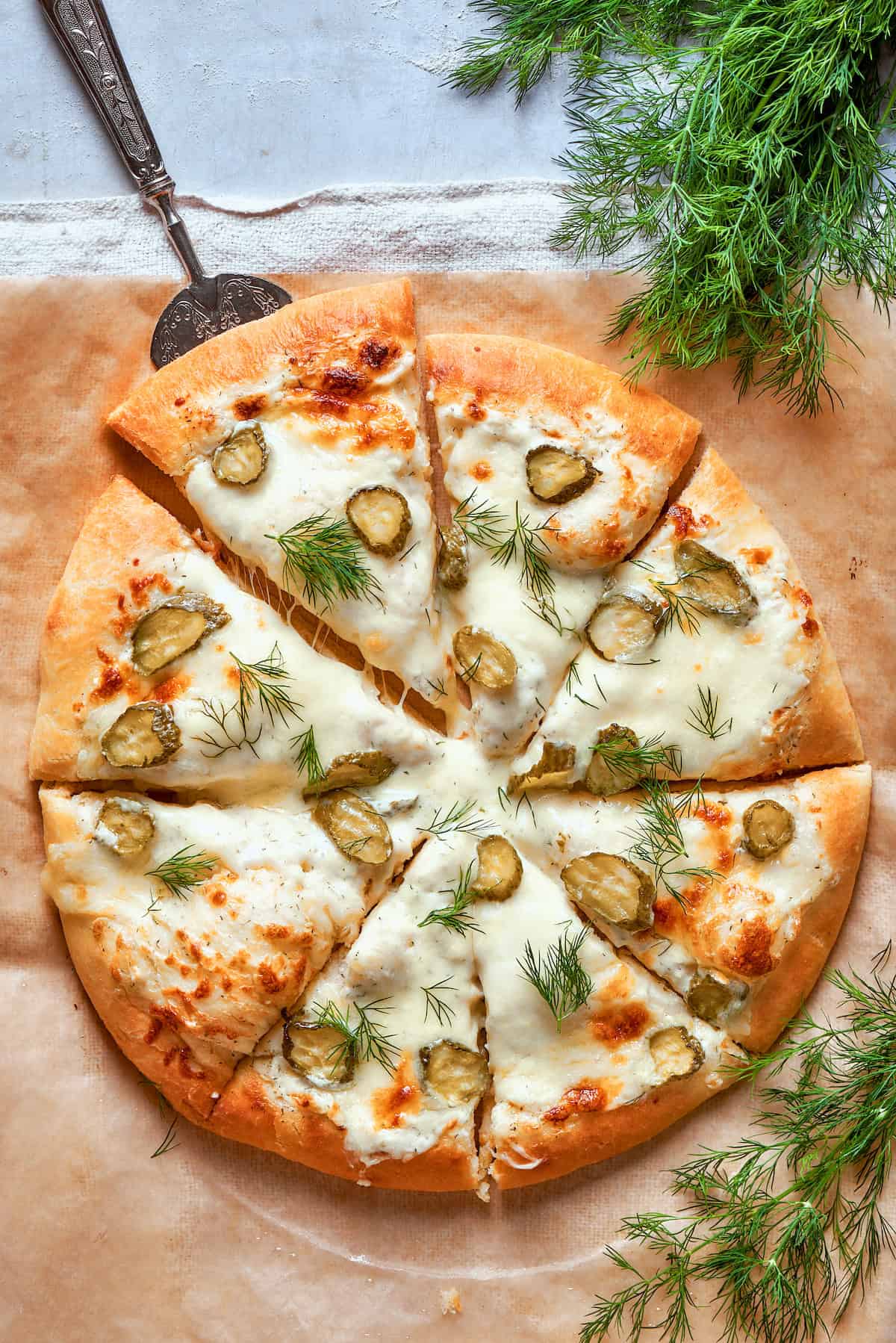 A whole pickle pizza shows it topped with white sauce, pickles, and dill.