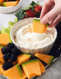A hand dips a piece of melon into fruit dip.