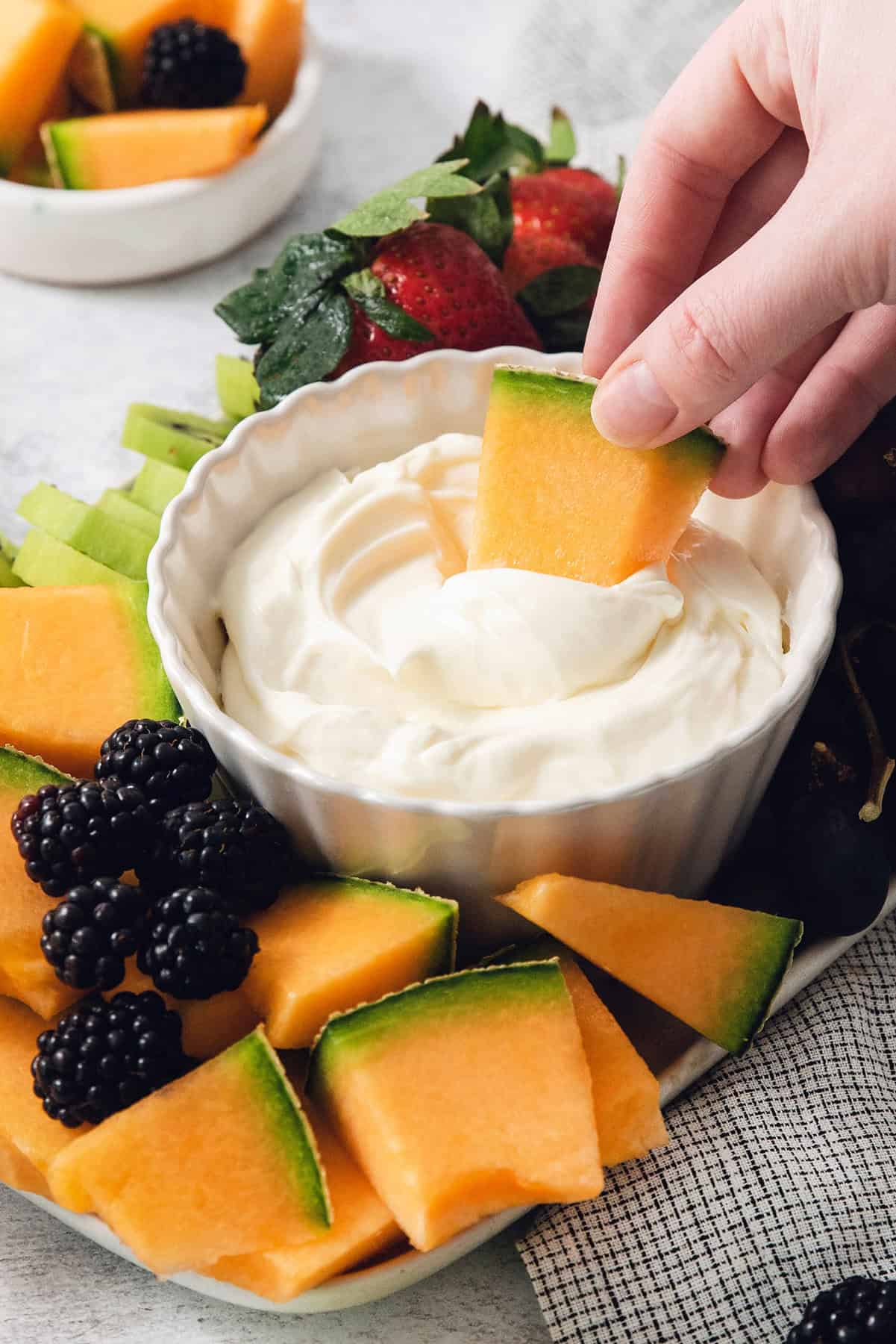 This 2-ingredient Fruit Dip is the perfect fluffy and creamy dessert dip featuring tangy cream cheese swirled with marshmallow creme. A hand dips a piece of melon into fruit dip.