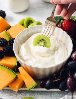 A fork dips a slice of kiwi into a bowl of fruit dip.