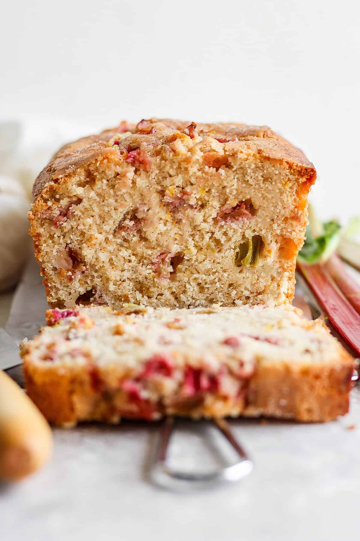This Rhubarb Bread is gorgeously moist and tender, warmly spiced with cinnamon and spiked with orange zest. A loaf of rhubarb bread cut to show a slice and the pink spots of rhubarb.
