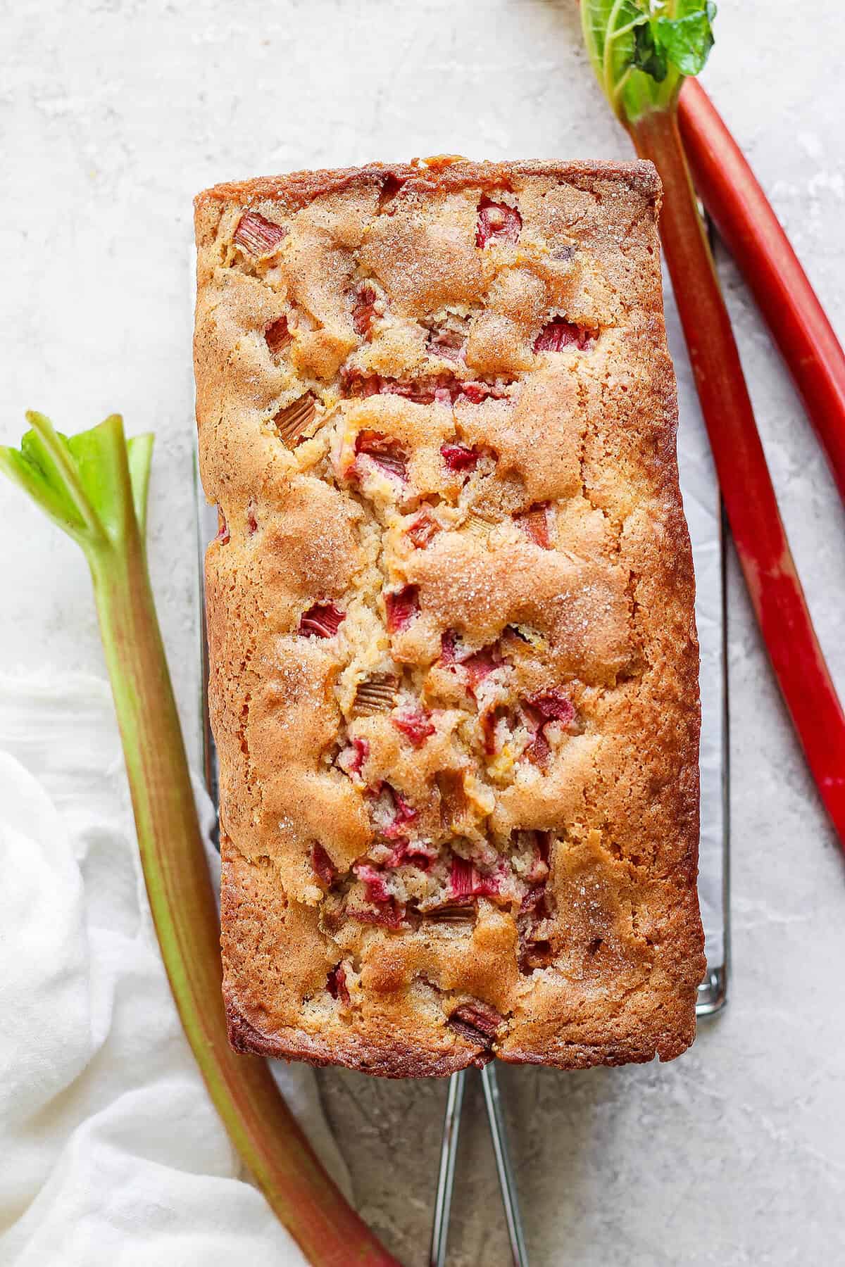 This Rhubarb Bread is gorgeously moist and tender, warmly spiced with cinnamon and spiked with orange zest. A baked rhubarb bread loaf with stalks of rhubarb next to it.