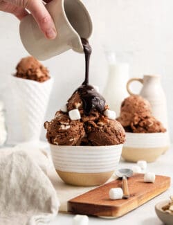Chocolate sauce is poured over scoops of rocky road ice cream in a bowl.