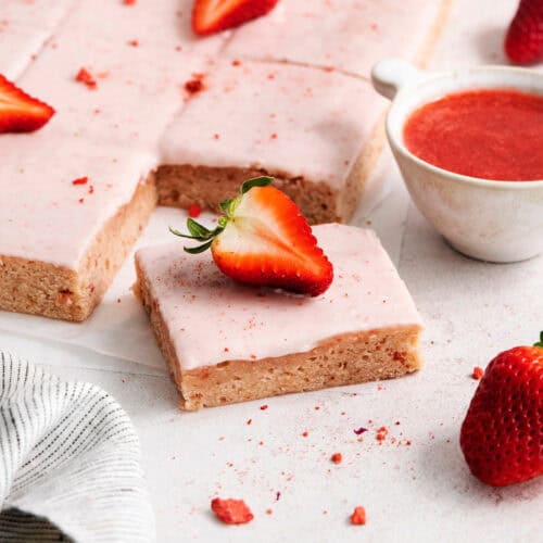 Pink icing-topped strawberry brownies are cut into squares, with strawberries scattered around them.