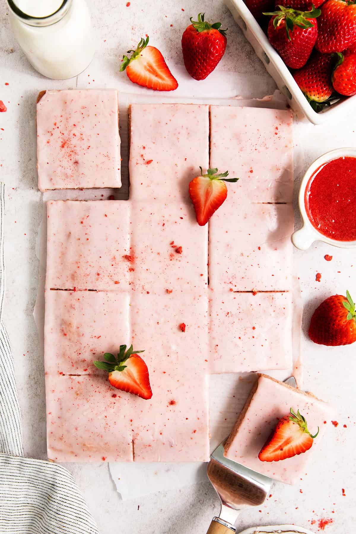 Pink icing-topped strawberry brownies are cut into squares, with strawberries scattered around them.