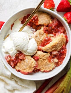 individual serving of strawberry rhubarb cobbler in a white bowl, with scoops of vanilla ice cream
