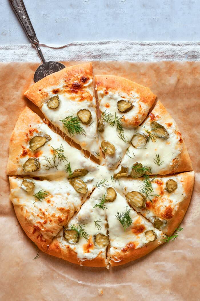 Creamy, crunchy, and puckery dill Pickle Pizza is a fun twist on classic pizza with white sauce, fresh and shredded mozzarella, dill, and plenty of pickles. overhead photo of a whole pickle pizza cut into individual pieces