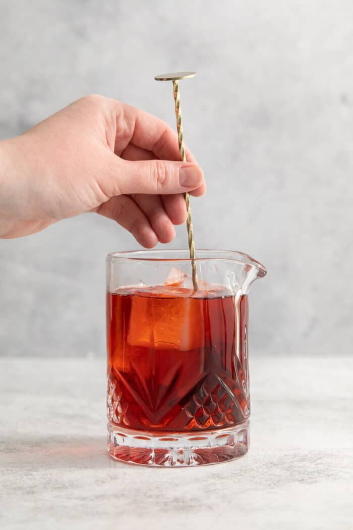 A hand uses a cocktail stirrer to stir a Negroni.