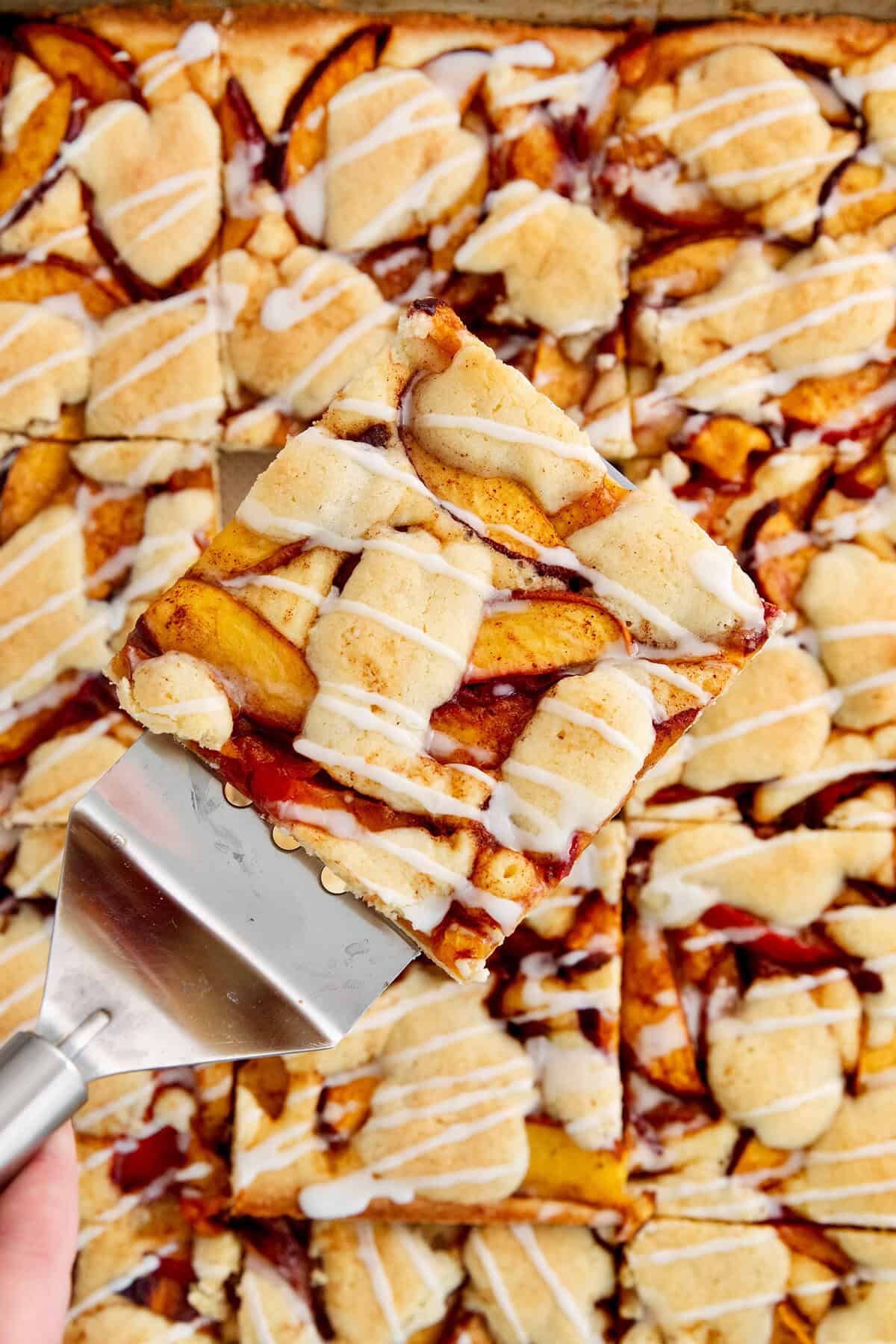 These Peach Bars feature a tender, buttery base topped with ripe, juicy peaches swirled with a touch of cinnamon. A serving spatula lifts out a peach bar.