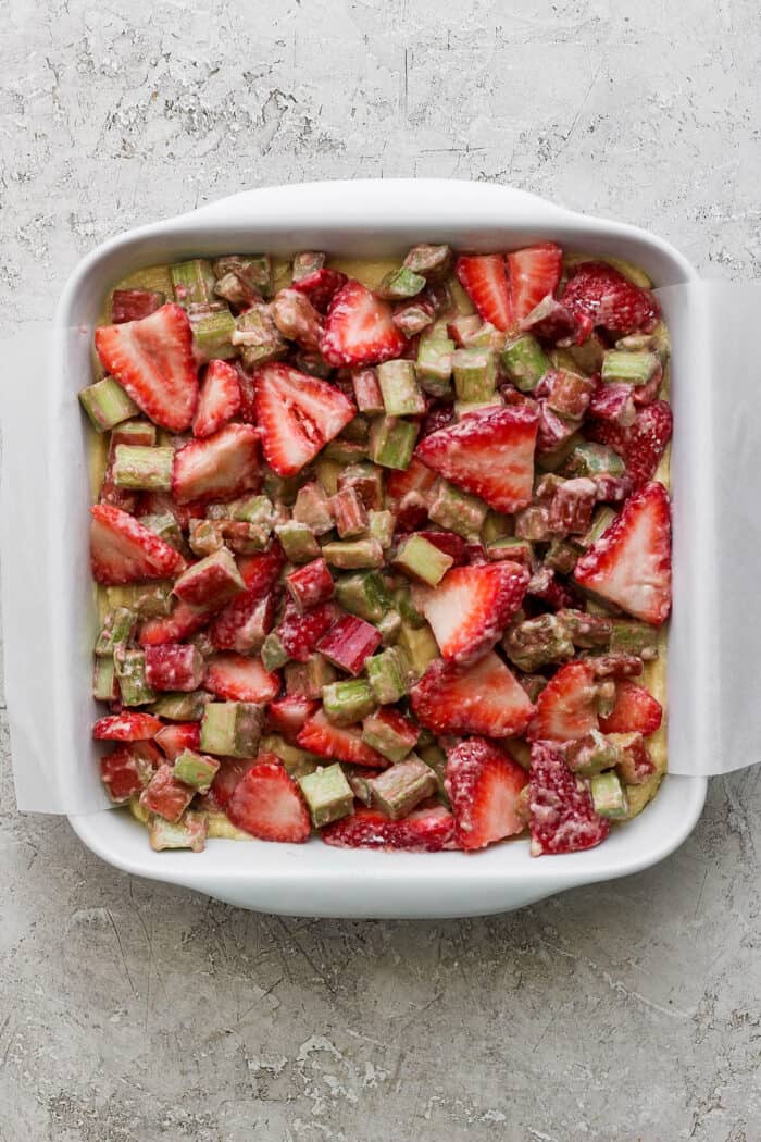 a square white baking dish with batter covered by a mixture of strawberries and rhubarb