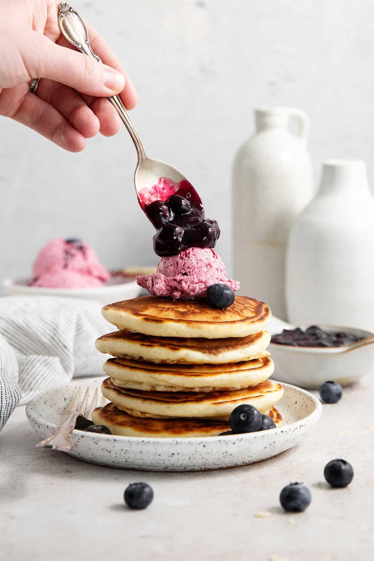This Blueberry Butter is lightly sweet and luxuriously fluffy, with an easy homemade fresh blueberry sauce as its base. A hand spoons blueberries on top of a scoop of blueberry butter on top of a stack of pancakes.