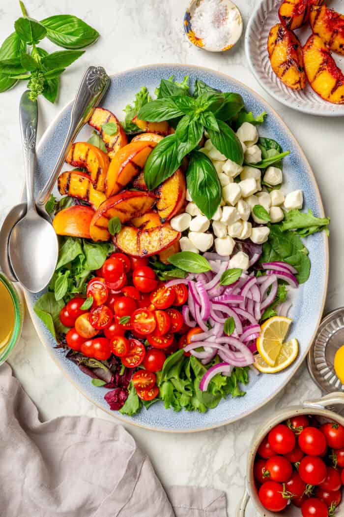 Sweet and juicy Grilled Peach Salad features lightly grilled peaches served with greens, cherry tomatoes, and fresh mozzarella. Salad greens are topped with grilled peach slices, red onion, fresh mozzarella, cherry tomatees, and basil leaves.