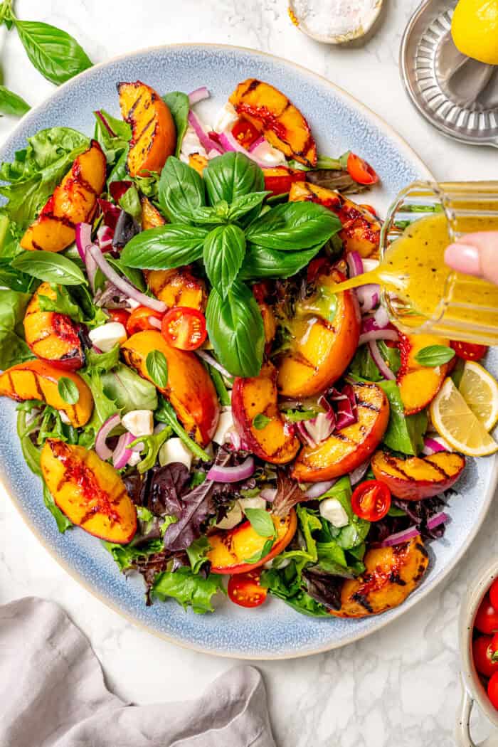 Sweet and juicy Grilled Peach Salad features lightly grilled peaches served with greens, cherry tomatoes, and fresh mozzarella. Dressing is poured over a serving platter of grilled peach salad.