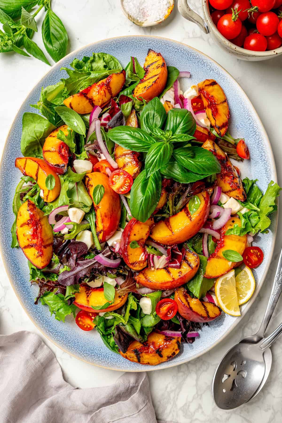 Sweet and juicy Grilled Peach Salad features lightly grilled peaches served with greens, cherry tomatoes, and fresh mozzarella. Basil leaves top a colorful serving platter of grilled peach salad.
