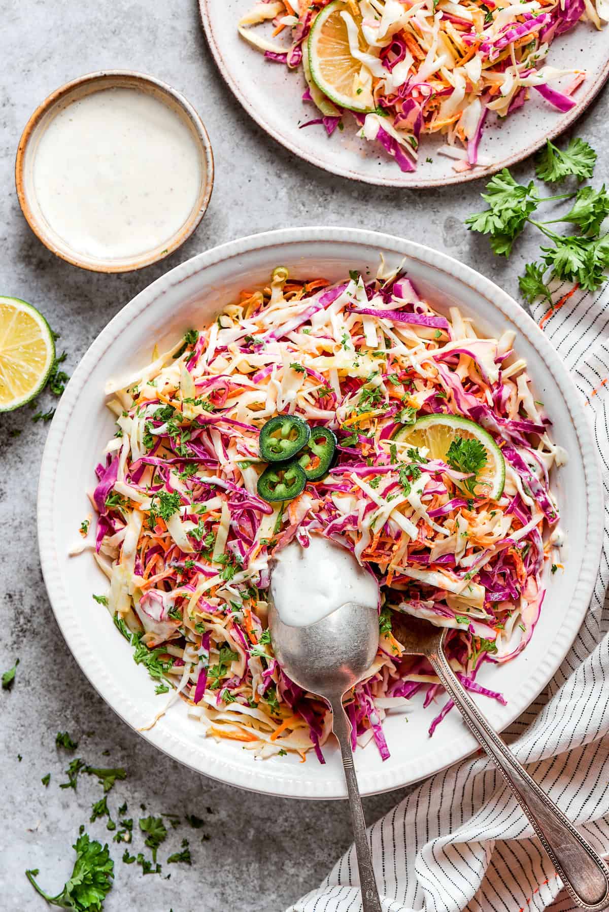 This jalapeño slaw recipe features a crunchy mix of vegetables including cabbage, cilantro, and carrots tossed in a lightly sweet dressing. Plates of jalapeño slaw with limes and salt around them.