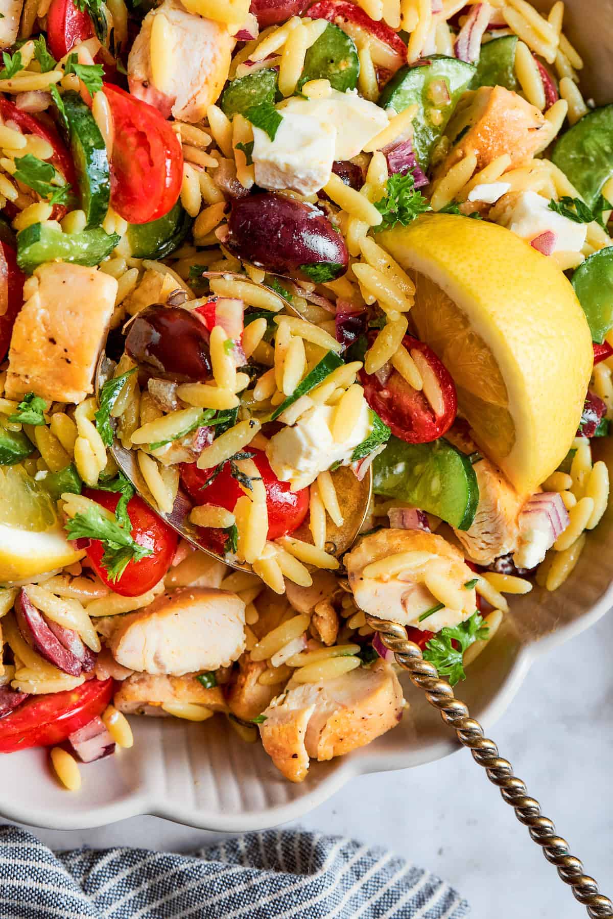 This easy Lemon Chicken Orzo Salad features fresh veggies, chicken, pasta, and a tangy lemon vinaigrette for a light yet filling meal. a serving spoon in a white bowl full of orzo salad