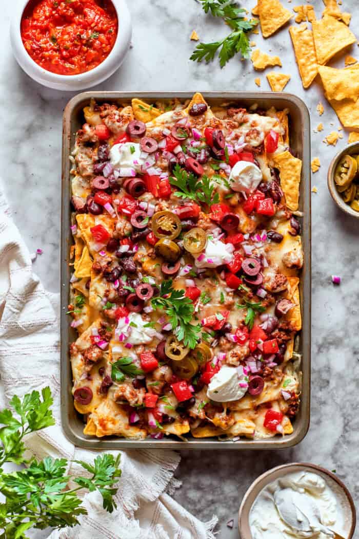 This Macho Nachos recipe features layers of crispy tortilla chips, melty cheese, and seasoned beef. Perfect for any casual gathering! A baked tray of macho nachos surrounded with bowls of sasa, sour cream, and more chips.