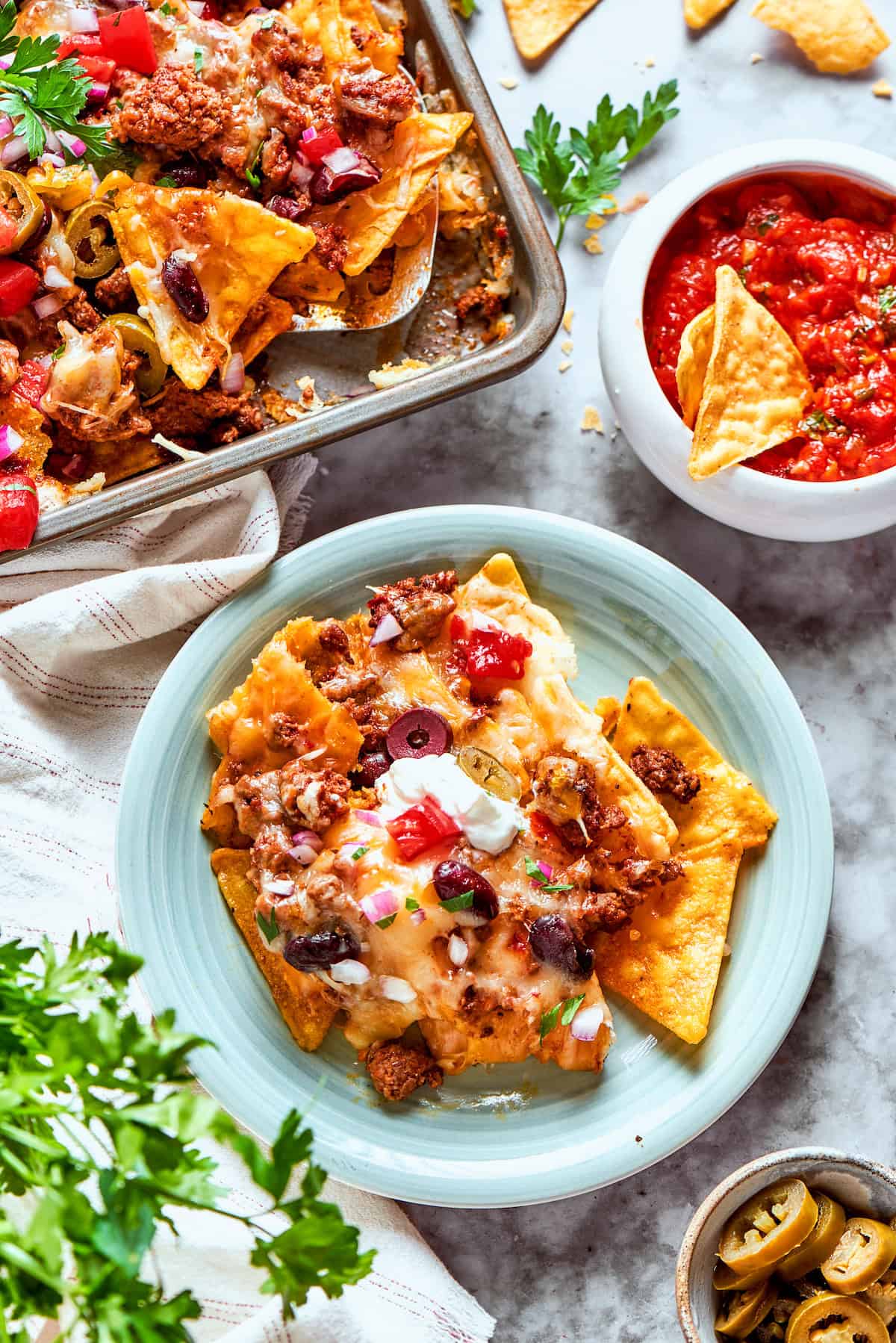 This Macho Nachos recipe features layers of crispy tortilla chips, melty cheese, and seasoned beef. Perfect for any casual gathering! A plate of macho nachos next to a tray of nachos and a bowl of salsa.