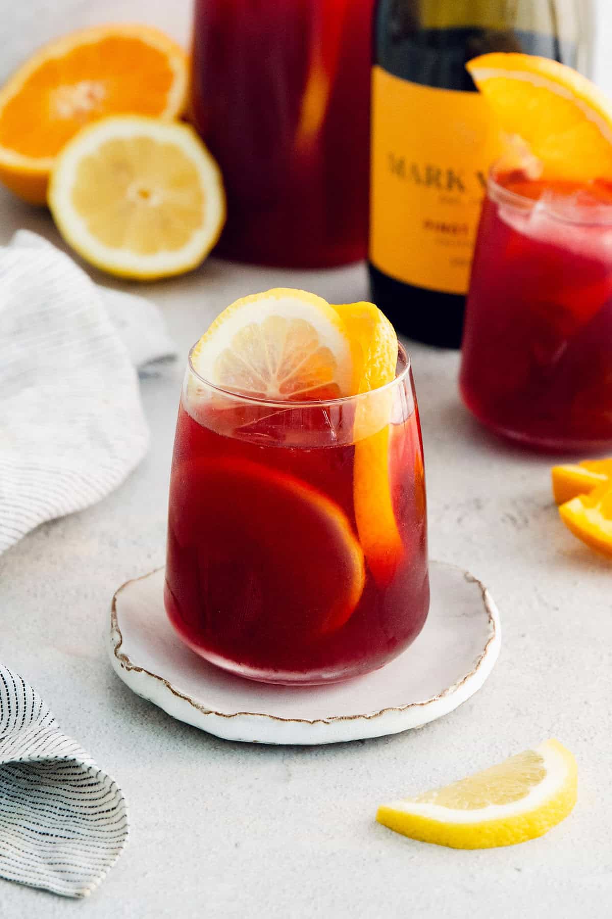 Tinto de Verano, a Spanish summertime cocktail, features fruity red wine, lemon soda, and red vermouth served over ice. A glass of red tinto de verano is served on a white plate with more glasses behind it.