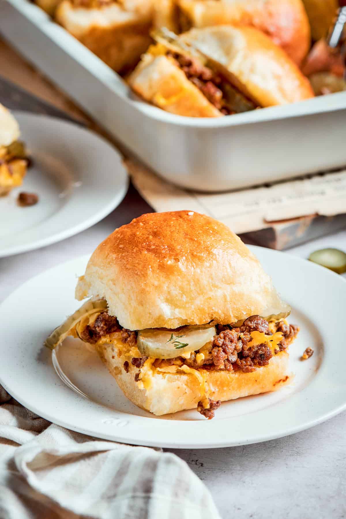 Homemade Cheeseburger Sliders star tender ground beef topped with plenty of melted cheddar cheese served on sweet rolls. A cheeseburger slider on a white plate with a pan of sliders in the background.