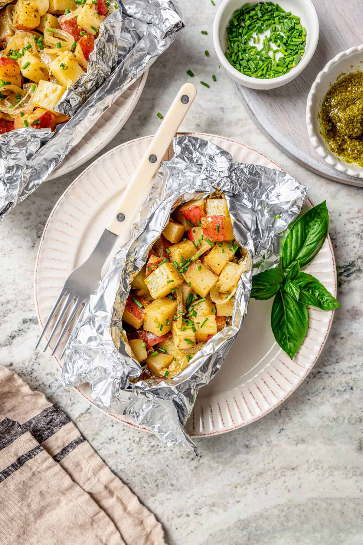 Savory Grilled Potato Packets feature grilled potatoes in foil dressed in tangy, garlicky pesto-ranch sauce and ready in less than 30 minutes. Grilled potato packets are served on plates with fresh basil leaves.