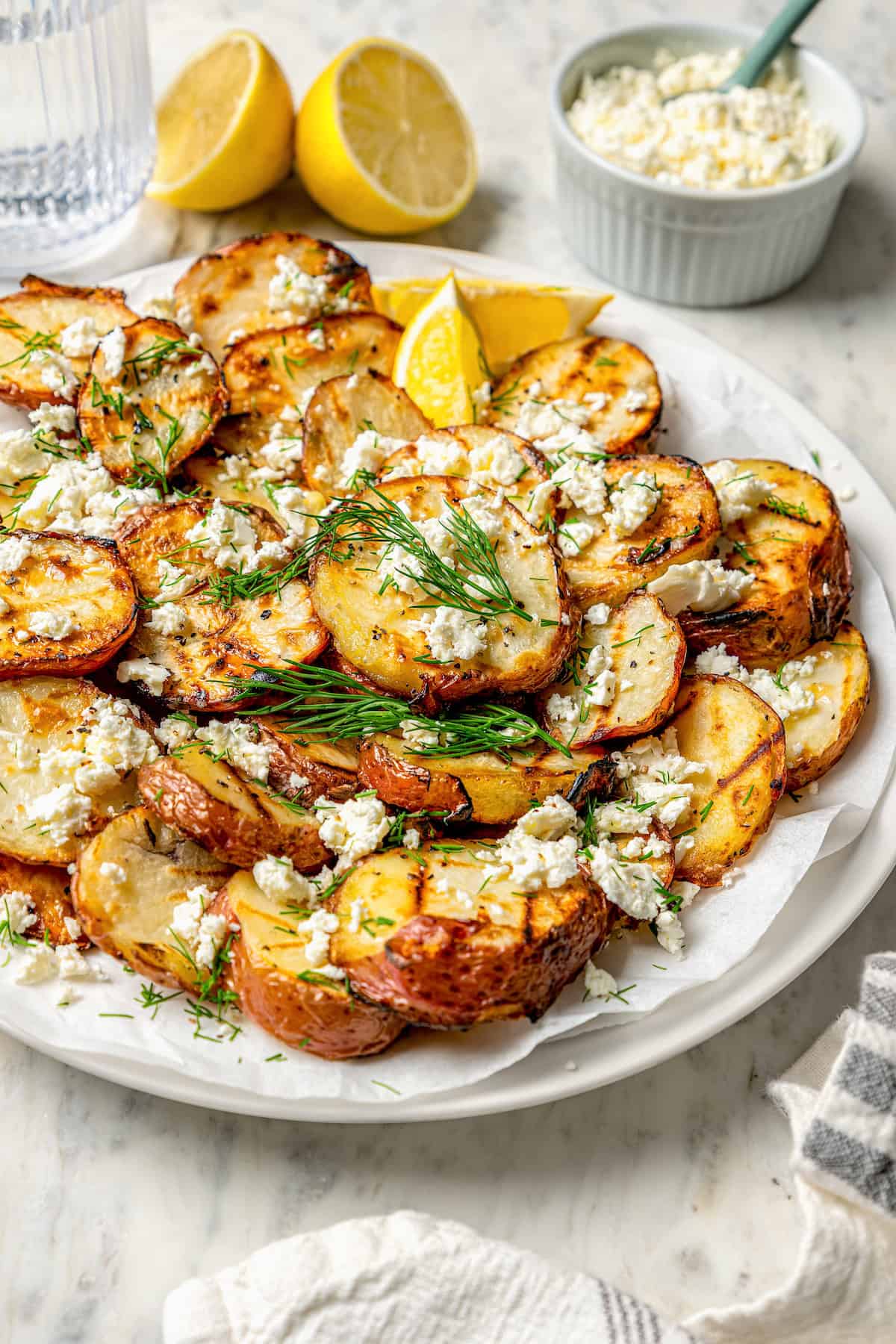Crisp-tender Grilled Red Potatoes are spiked with the brisk flavors of dill, feta cheese, and lemon and grilled to perfection. Lemons and a bowl of cheese are in the background with a plate of grilled red potatoes in front of them.