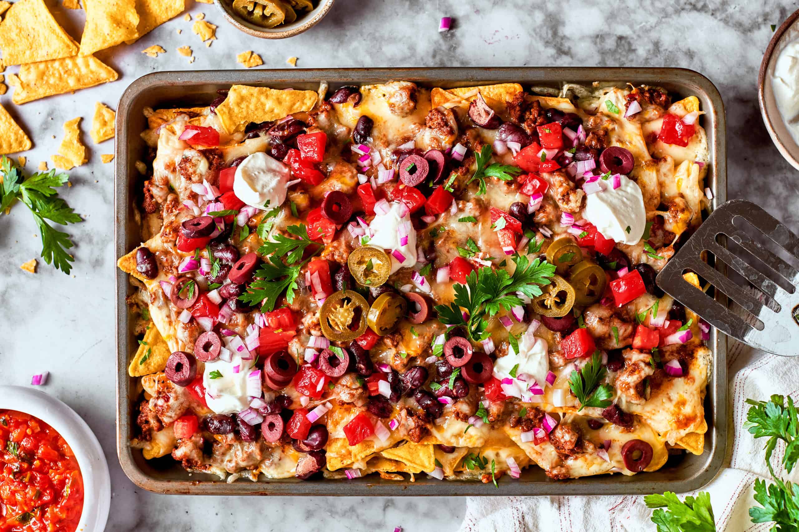 This Macho Nachos recipe features layers of crispy tortilla chips, melty cheese, and seasoned beef. Perfect for any casual gathering! overhead photo of a rimmed pan full of baked cheesy nachos loaded with meat and vegetables
