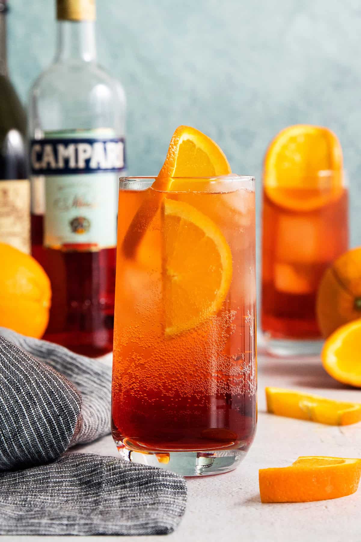 This 3-ingredient Americano Cocktail is made from equal parts Campari and sweet vermouth plus a splash of bubbly club soda. A bottle of Campari stands in the background with two Americano cocktails.