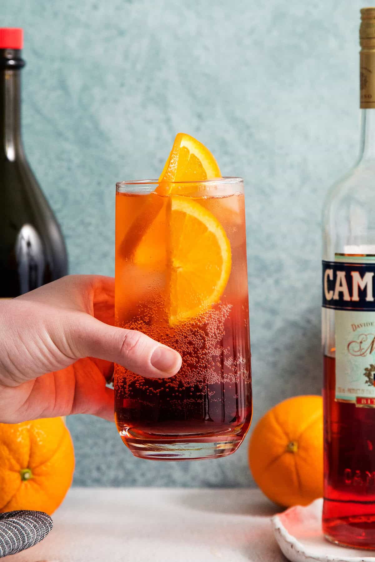 This 3-ingredient Americano Cocktail is made from equal parts Campari and sweet vermouth plus a splash of bubbly club soda. A hand holds a tall glass of Americano cocktail garnished with orange slices.
