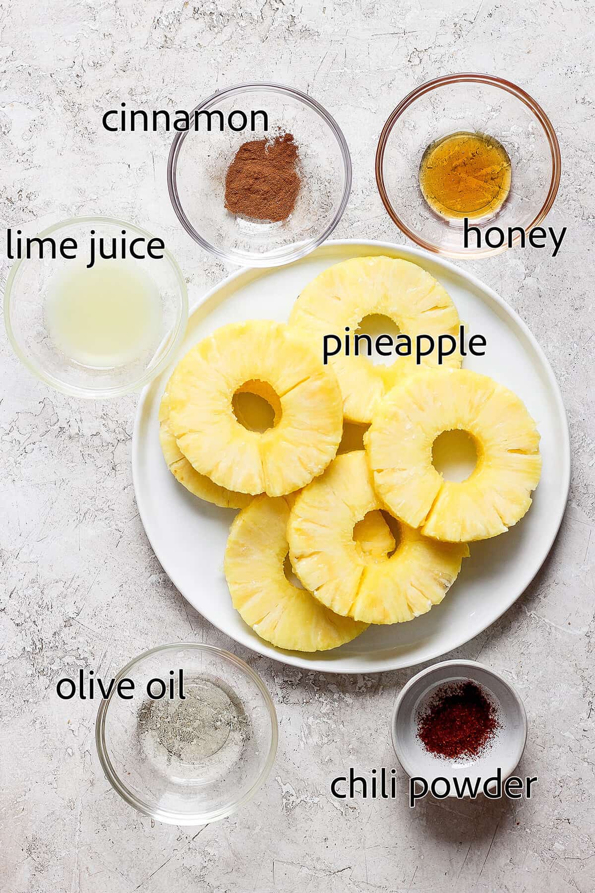 Lightly caramelized and so juicy Grilled Pineapple is a blend of sweet and spicy, perfect on top of burgers or as a dessert. Ingredients for grilled pineapple are labeled: pineapple, honey, oil, lime juice, chili powder, cinnamon.