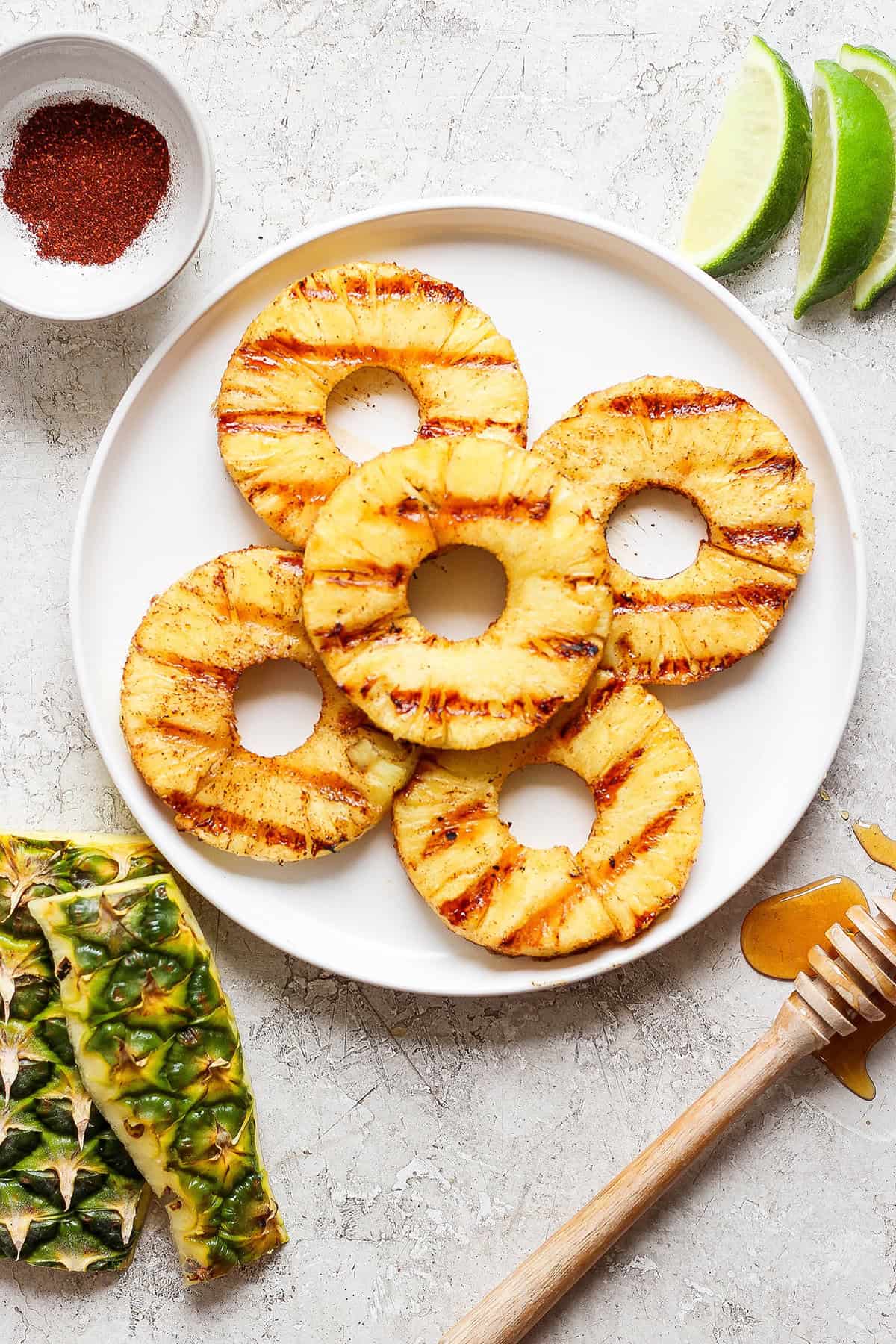 Lightly caramelized and so juicy Grilled Pineapple is a blend of sweet and spicy, perfect on top of burgers or as a dessert. Five rings of grilled pineapple on a white plate.