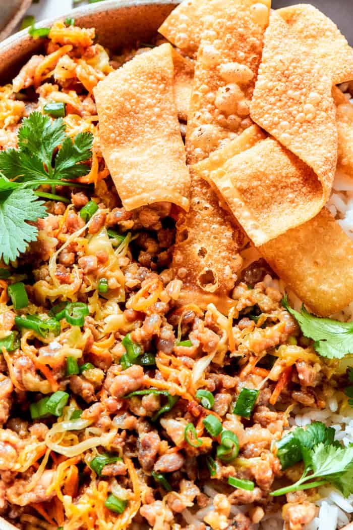 This easy Egg Roll in a Bowl recipe is loaded with ground pork, cabbage and carrots. Served with crispy wonton strips and a tasty Asian sauce! close-up photo of finished egg roll filling served over white rice, with wonton chips
