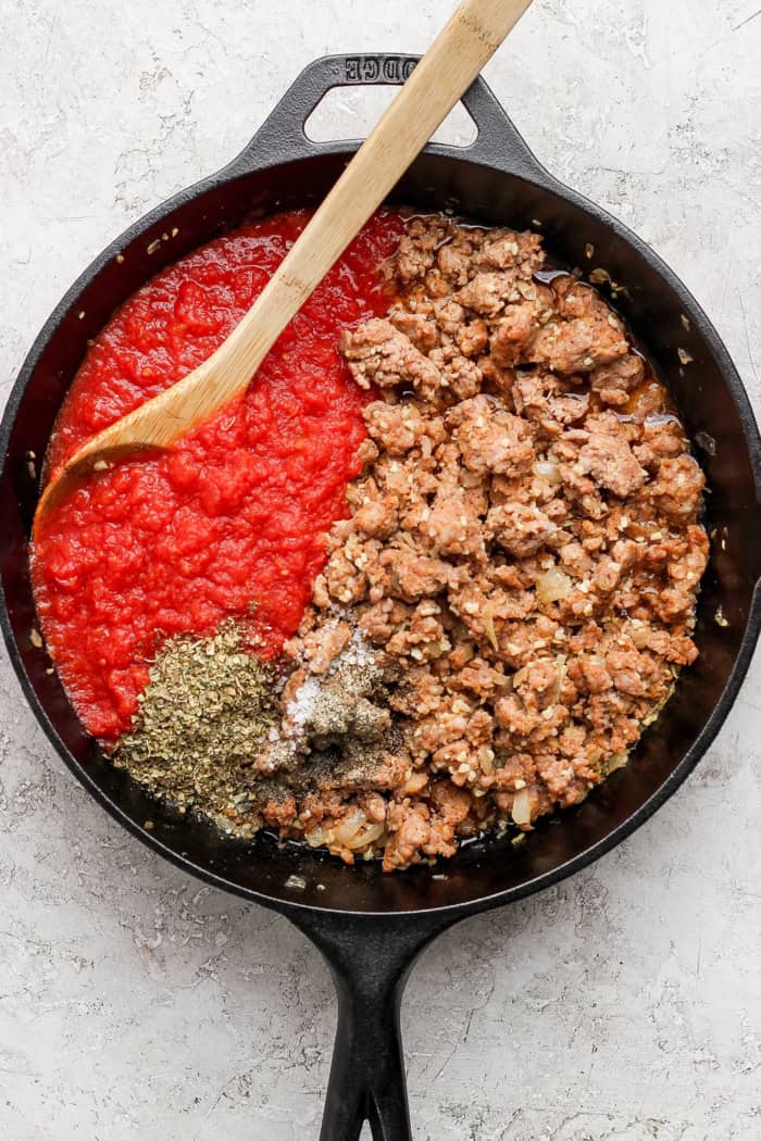This Lasagna Dip has melty pockets of cheese, meaty sausage, and zesty tomato sauce, all ready for scooping with garlic bread dippers. YUM! Overhead view of sausage, seasonings, and crushed tomatoes in skillet before stirring