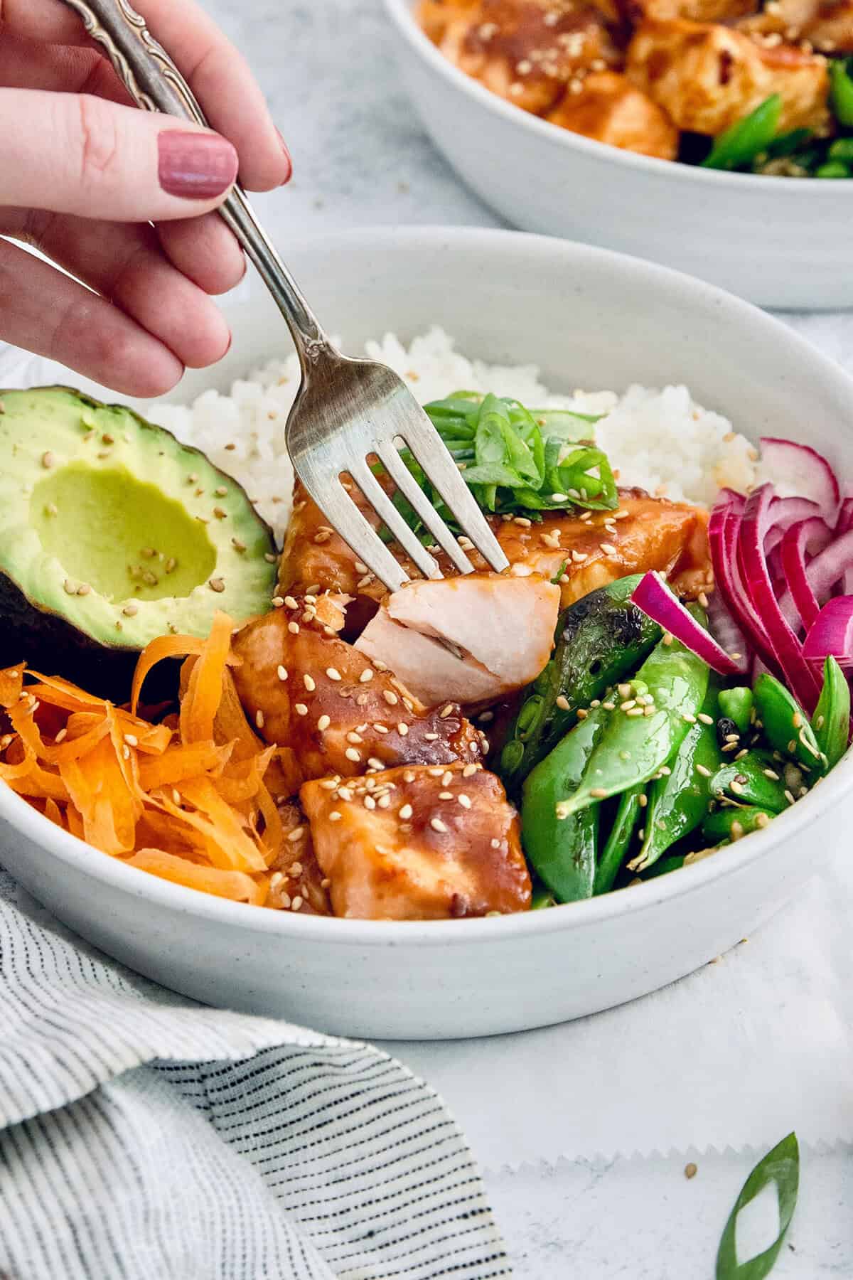 Teriyaki salmon bowls are an easy dinner ready in less than 20 minutes, with tender salmon, sticky teriyaki sauce, and lots of fresh veggies! Fork piercing teriyaki salmon bite in meal bowl