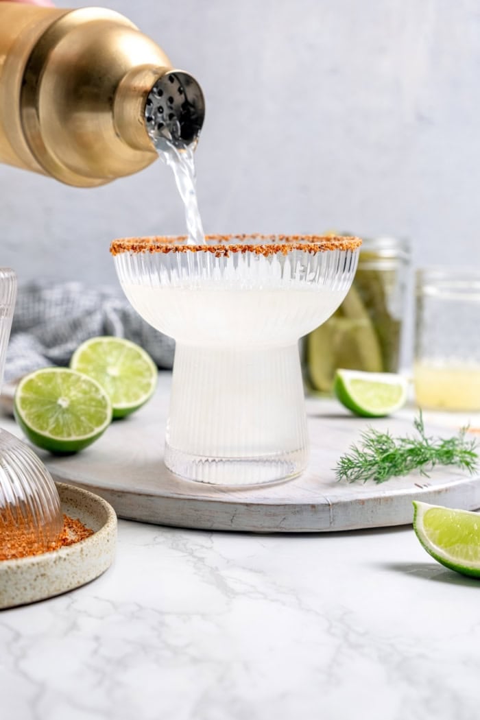 This Pickle Margarita recipe is bright and citrusy just like the classic, but it has a little extra pop of flavor to make it truly special! Pouring pickle margarita into glass with ice and Tajin rim